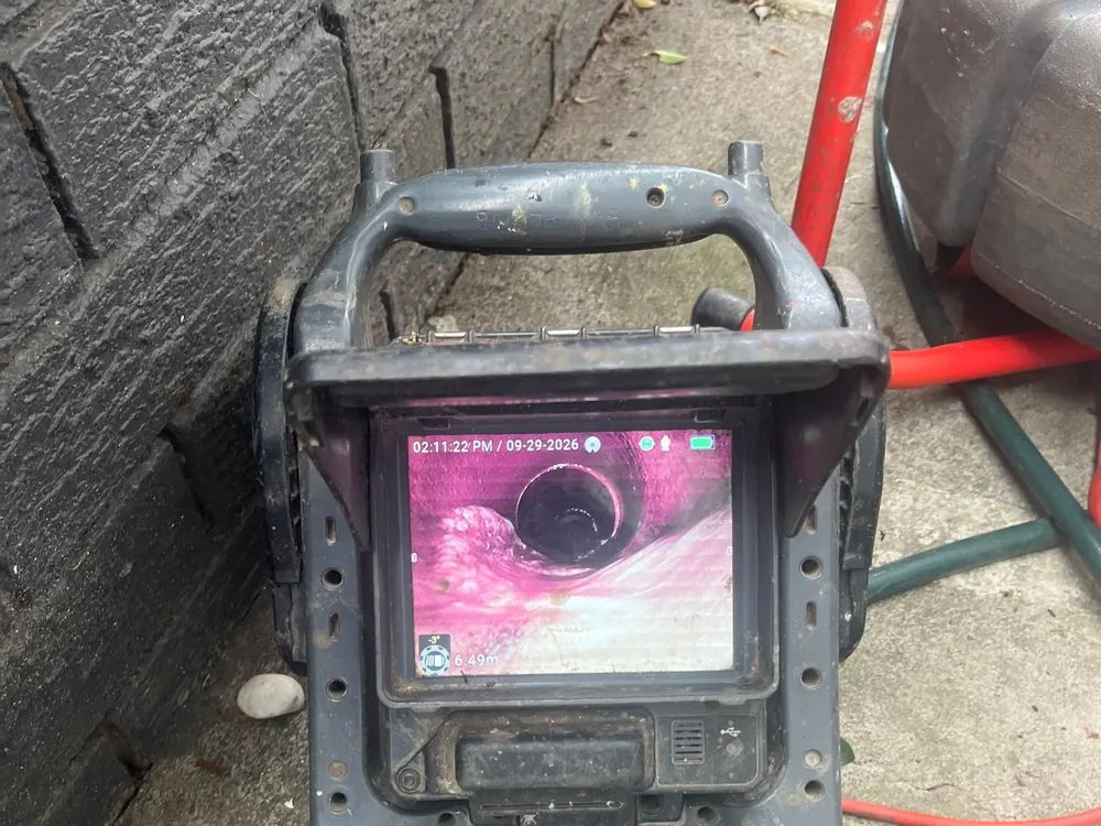 Drain camera monitor showing live pipe footage on the ground beside a house wall.